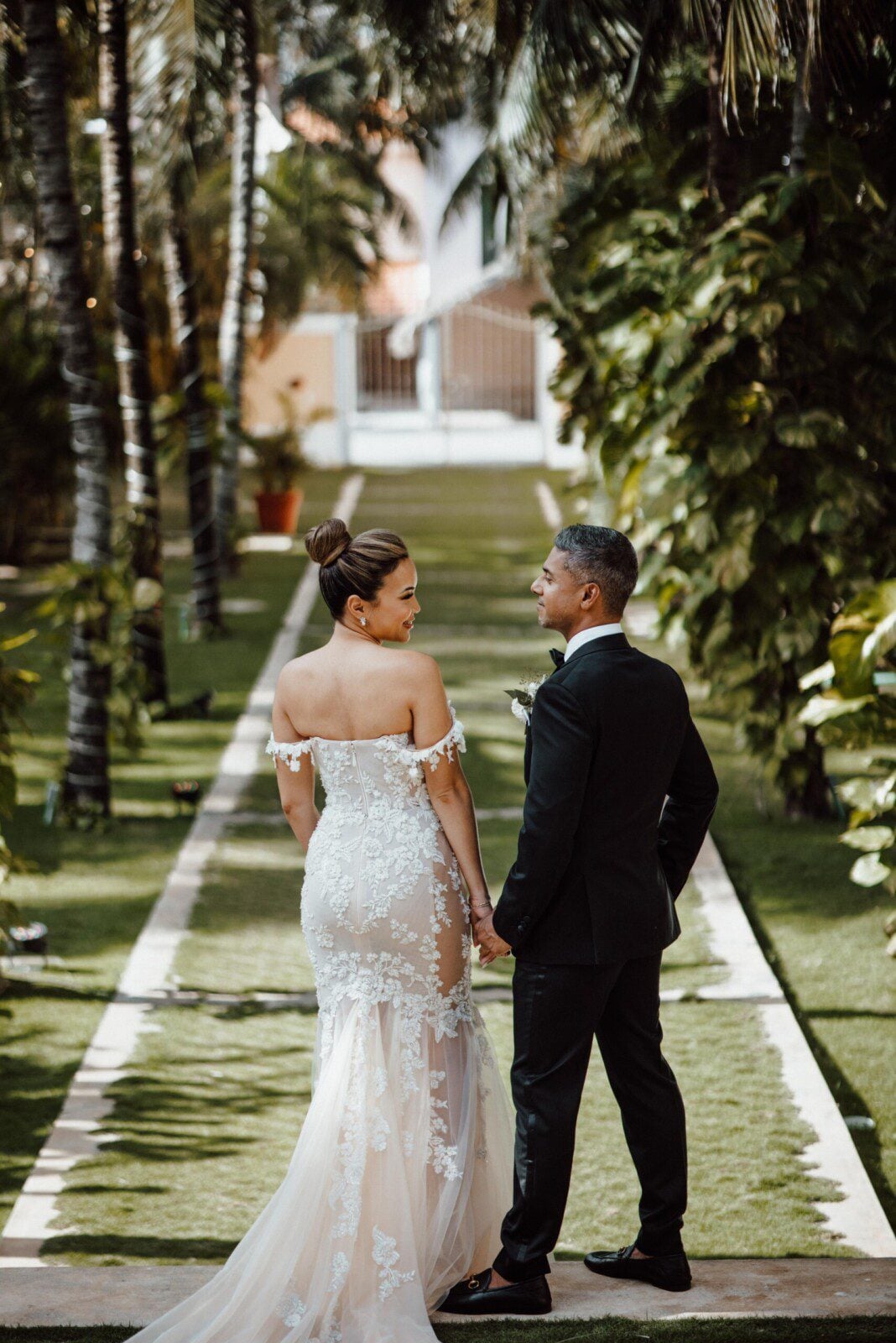 A bride and groom hold hands on a palm-lined garden path.