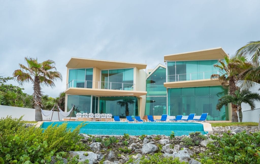 Villa Gauguin pool, blue loungers, and glass facade viewed from the rocky shore — Akumal, Mexico