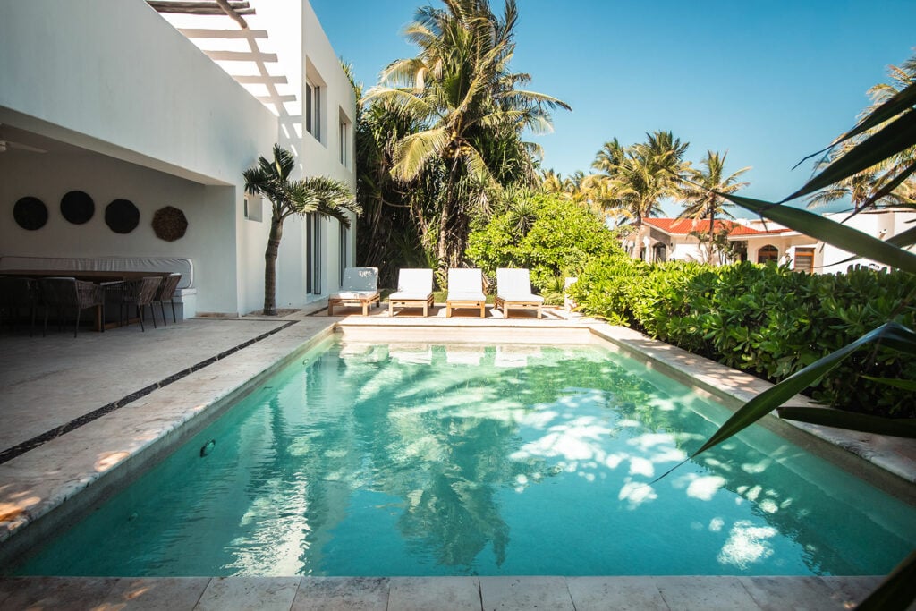 Casa Clara, Playacar, Riviera Maya: modern white villa with pool, lounge chairs, palm trees, and greenery under blue sky.