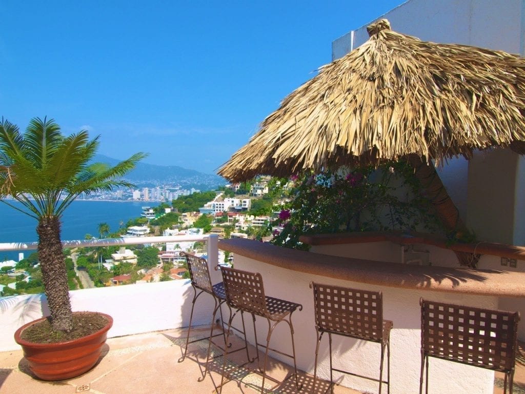Casa Cielito Lindo terrace bar beneath a thatched roof with a potted palm and bay view — Acapulco, Mexico