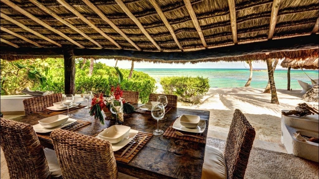 Puerto Estate dining table beneath a thatched roof facing palms and sea — Puerto Aventuras, Mexico