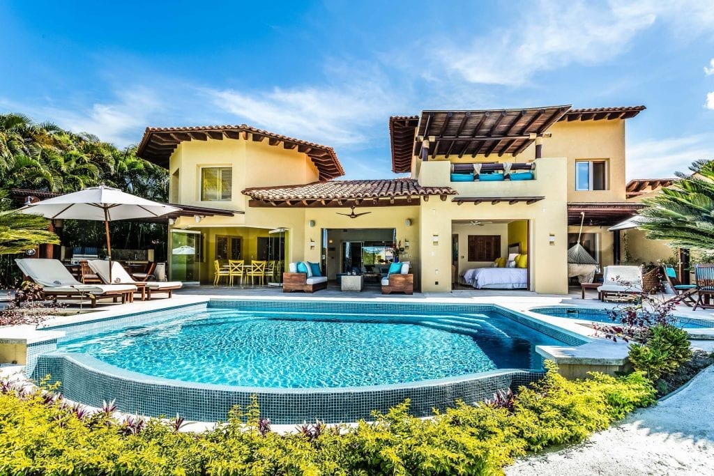 Villa Ipanema yellow exterior facade behind a curved pool and sun loungers — Punta Mita Nayarit, Mexico