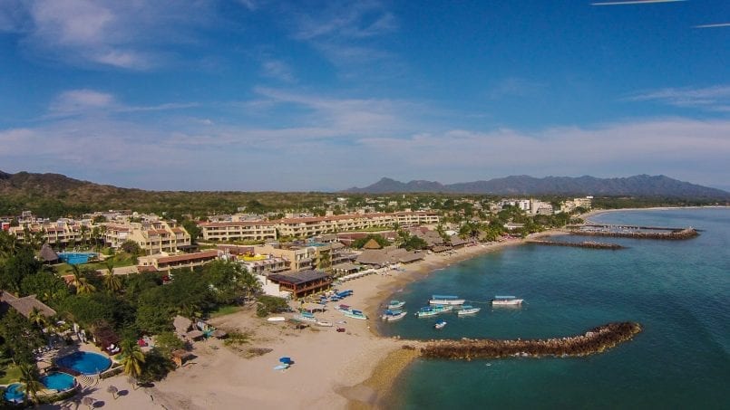 Early afternoon at El Anclote beach in Punta de Mita, capturing sunlit condos, bustling beachfront restaurants, anchored boats, and the sprawling sandy shore.