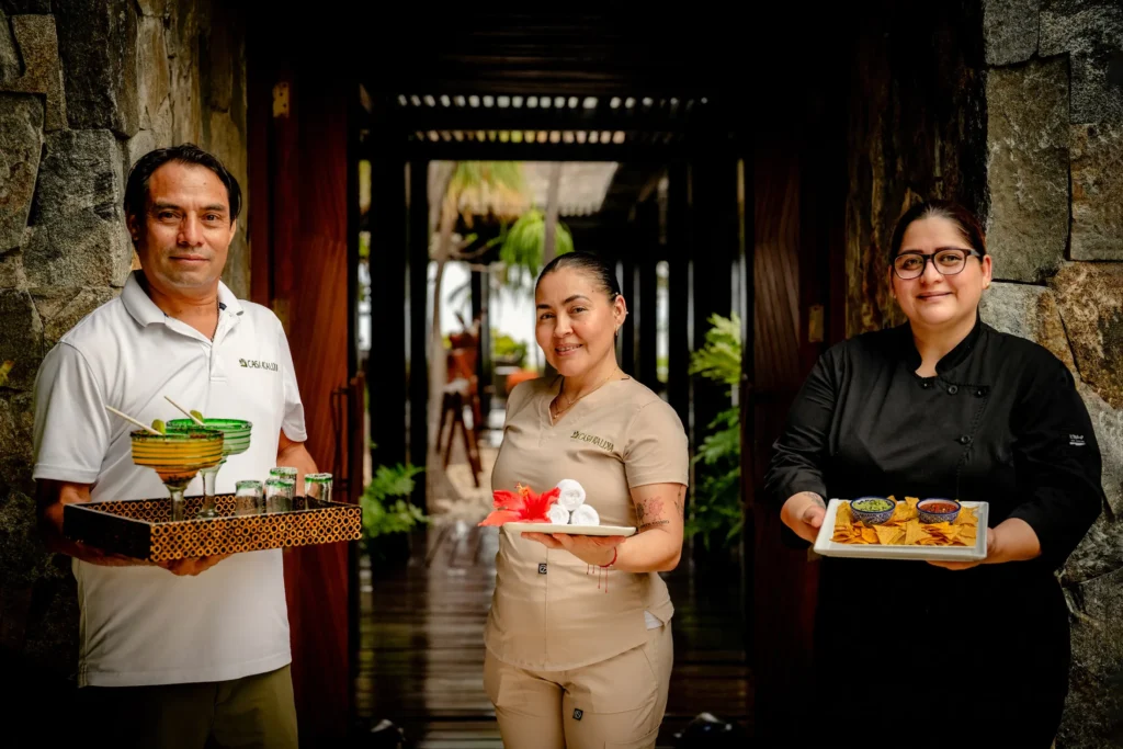 Three members of a villa staff at the door of a house, one with a tray of drinks, one with a cake, one in chef's whites with a platter