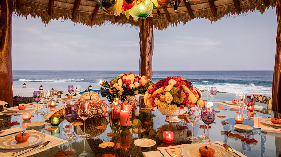 Elegant dinner table set on a luxury villa terrace in Mexico, overlooking a breathtaking ocean view, with high-end furnishings and festive decor under a sunset sky.