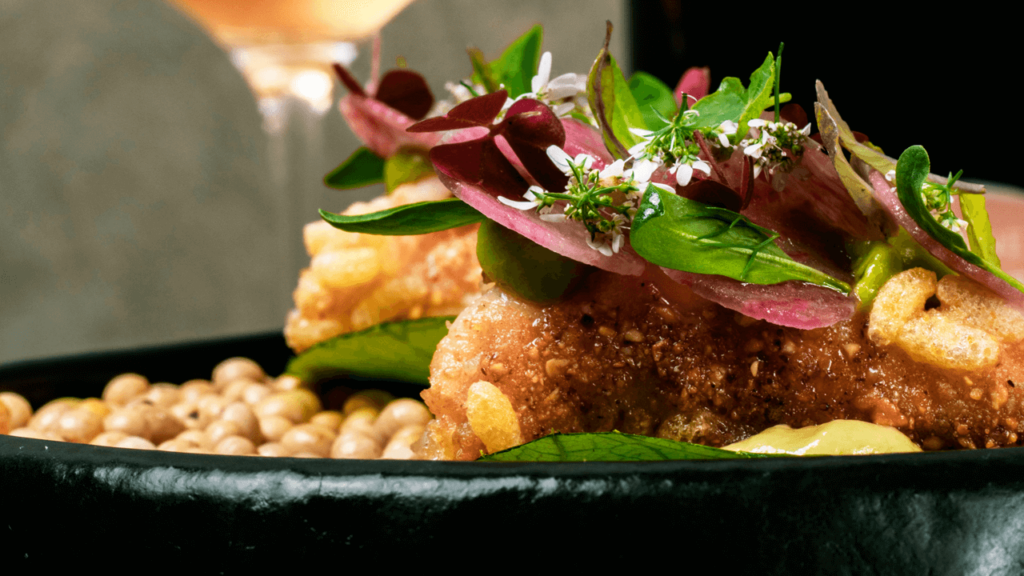 Close-up of a gourmet dish featuring crispy textures, fresh herbs, and vibrant garnishes, served on a black plate.