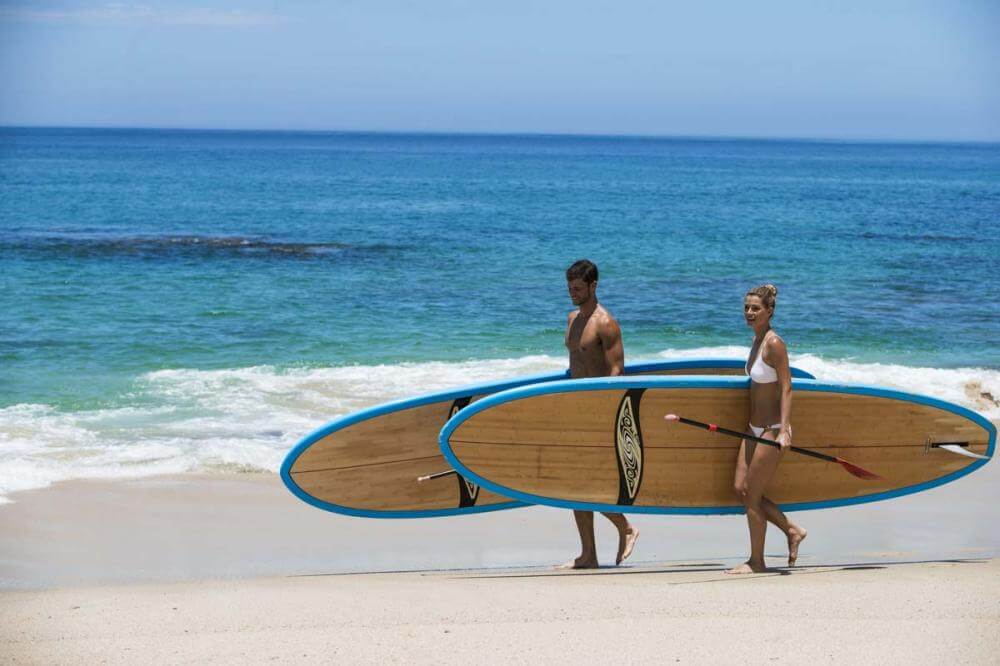 Enthusiastic individuals carrying stand up paddle boards along the pristine shoreline of Palmilla Beach, ready to embrace the serene azure waters.