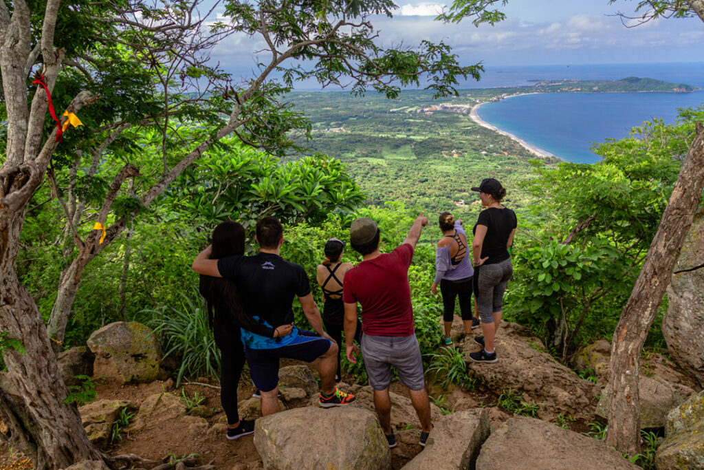 An image showing a group of people hiking on a scenic trail at El Cerro del Mono in Punta Mita, Mexico, with lush greenery and a panoramic view of the peninsula and ocean in the background.