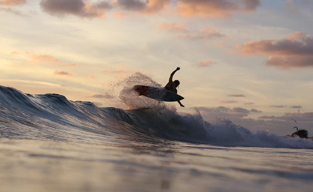 An image showing surfers riding waves during a stunning sunset at Punta Mita, Mexico, with vibrant hues of orange, pink, and purple reflecting on the ocean.