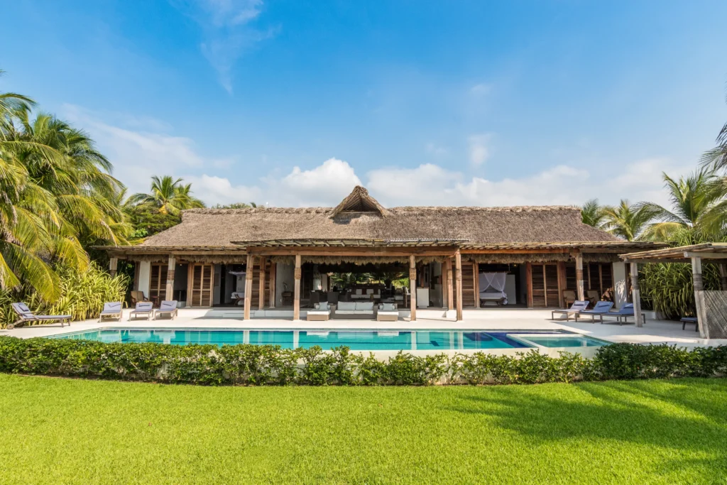 Casa Rhino thatched villa behind a pool and palm lawn under blue sky — Puerto Escondido, Mexico