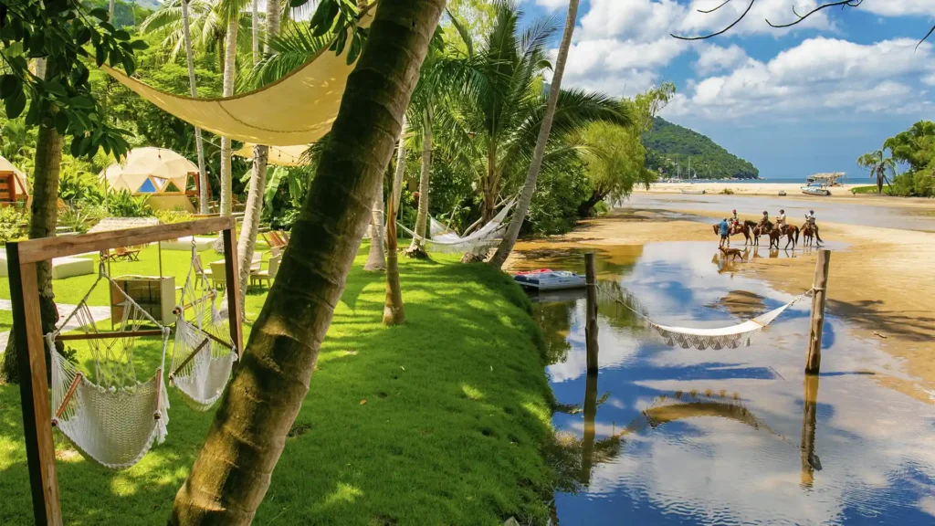 Hammocks by palm trees at Santuario Yelapa Mexico Wellness & Yoga Retreat, overlooking horses on the serene beach and lagoon.