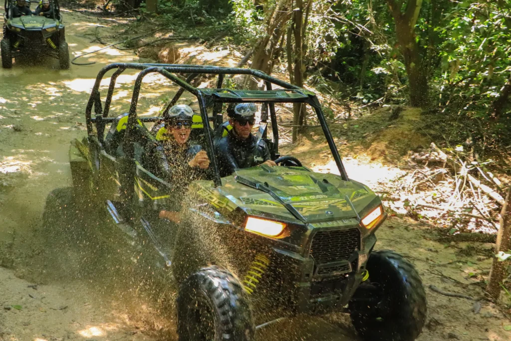 A buggy with headlights on pushes through a shallow creek under dense overhanging jungle.
