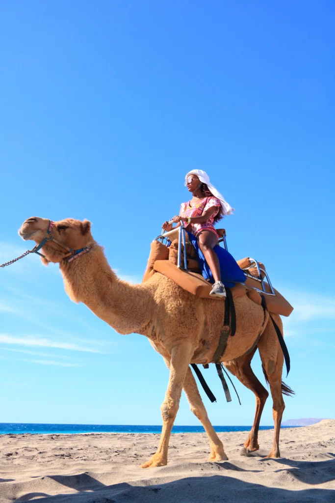 Two riders sit on a camel walking along the sand by the surf.
