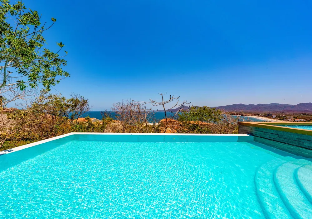 A turquoise pool edge over the bay under a clear blue sky, hills on the far shore