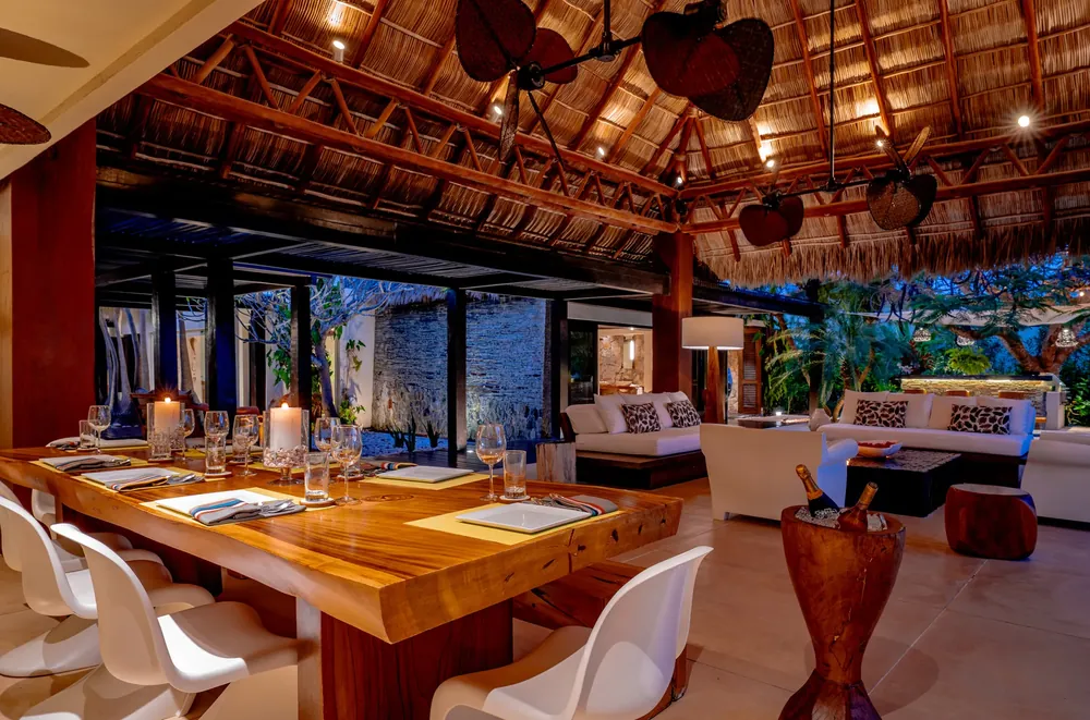 A long wooden dining table under a high palapa roof, open to the garden