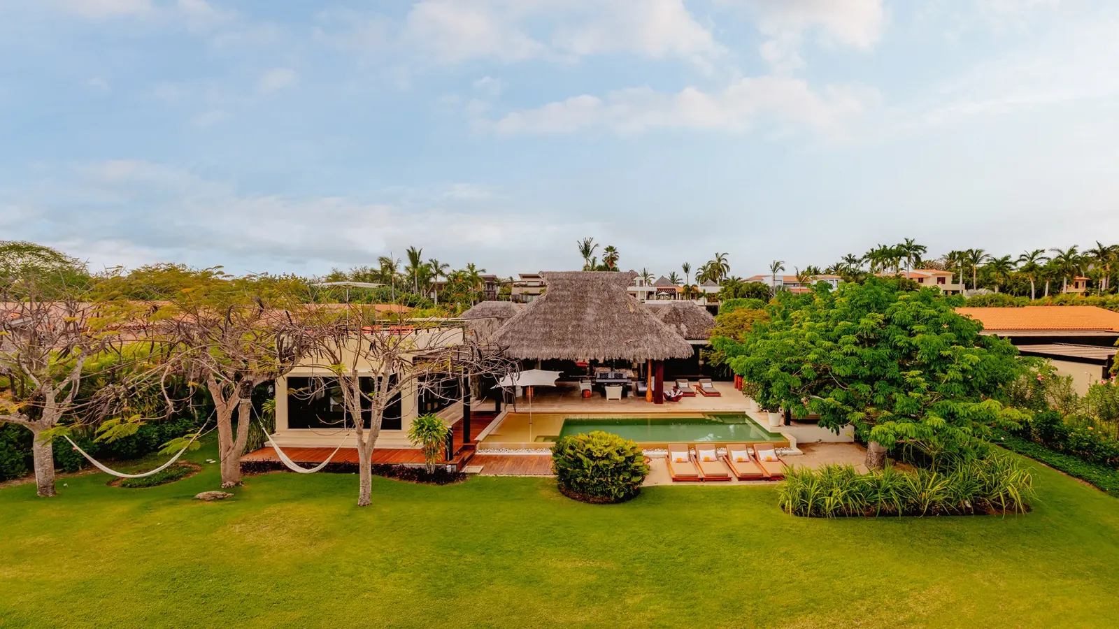 Palapa roofs of Casa Kalika behind a wide lawn and palms, in late afternoon light