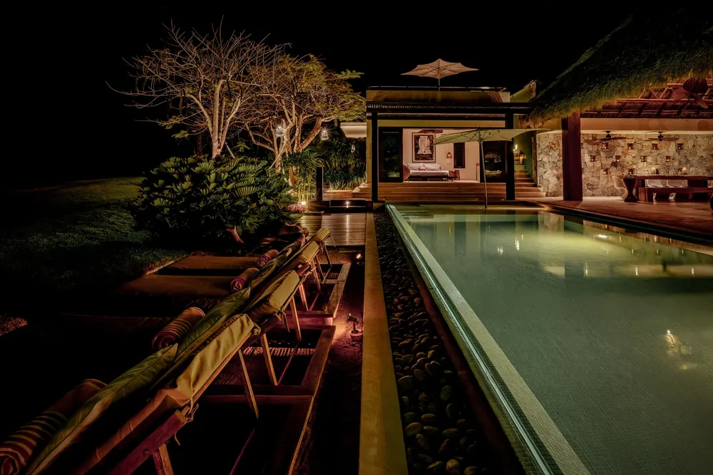 A long lit pool at night with loungers along its edge and trees lit from below