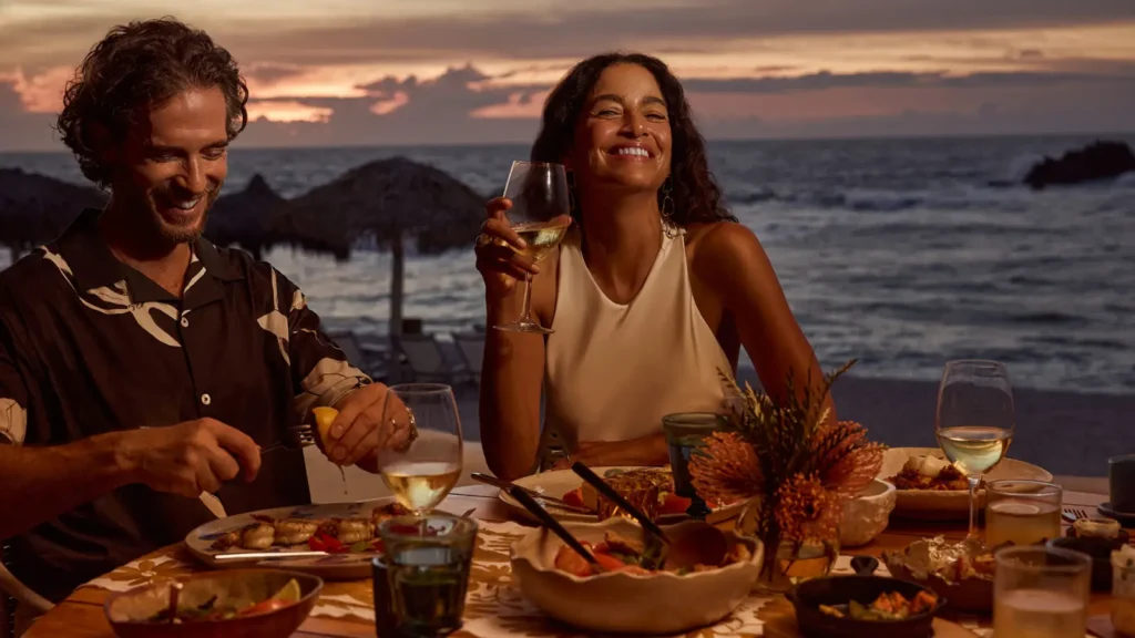 A man and a woman laughing over a seafood dinner at a table by the sea at sunset, Four Seasons Punta Mita
