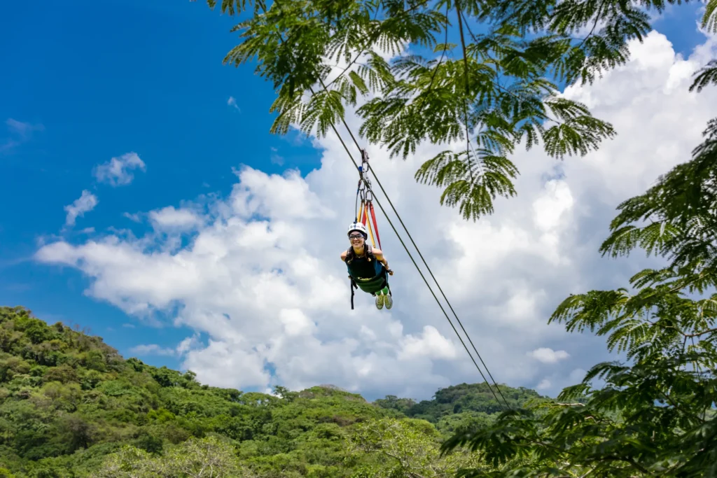 A rider hangs face-down from a long cable with white cloud and green hills behind.
