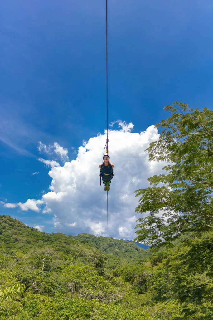 Seen from directly below, a rider travels a cable against tall white cloud and blue sky.