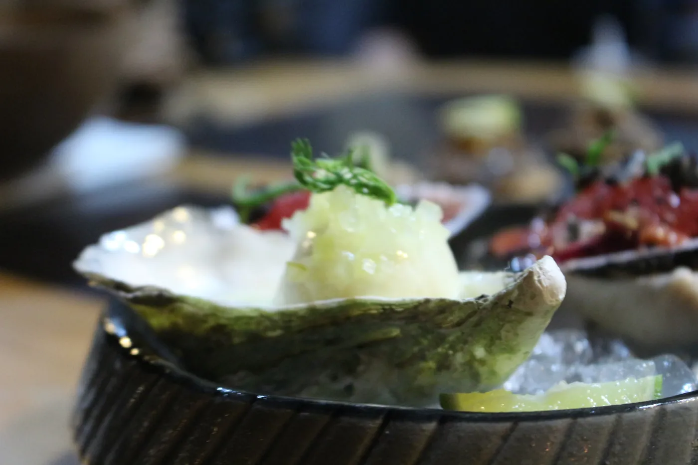 A close-up of an oyster topped with pale granita and a sprig of herbs on crushed ice with a lime wedge
