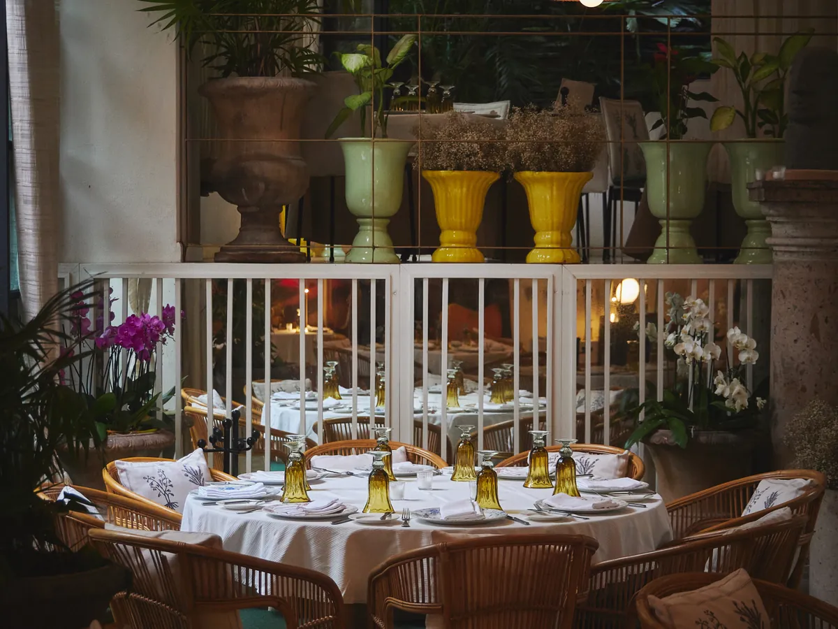 A round white-clothed table with rattan chairs and green glasses under a rattan lamp, with orchids and yellow urns on a railing behind