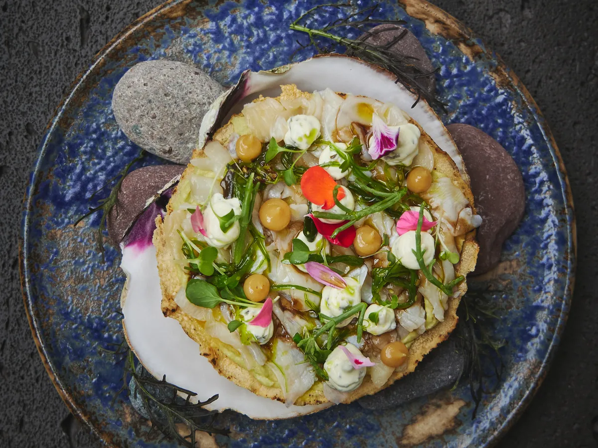 A round dish topped with green herbs, pink flowers and chickpeas on a blue ceramic plate on a dark stone table