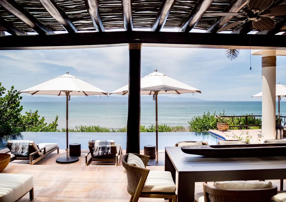 A covered terrace with a dark wood dining table, loungers and two white umbrellas beside the pool, with the sea beyond, Casa Tres Reinas, Punta de Mita