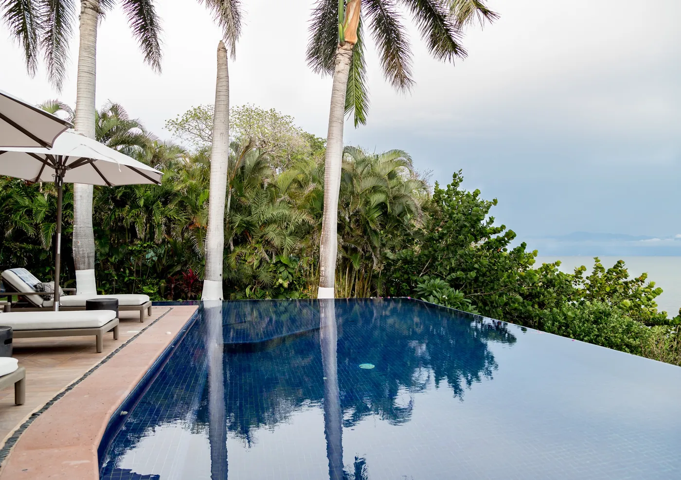 A dark-tiled infinity pool with loungers and a white umbrella under tall palms, with the sea and the far shore of the bay beyond, Casa Tres Reinas, Punta de Mita