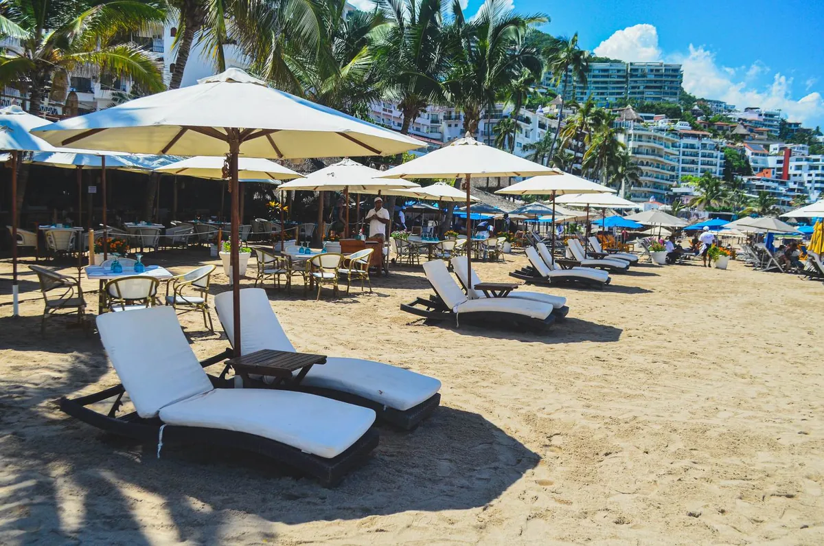 White loungers and umbrellas on the sand at a beach club, with palm trees and a hillside of white buildings behind