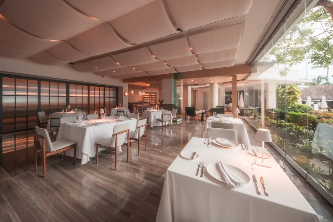 White-clothed tables and grey chairs under a draped white ceiling, with a glass wall looking onto palms and gardens