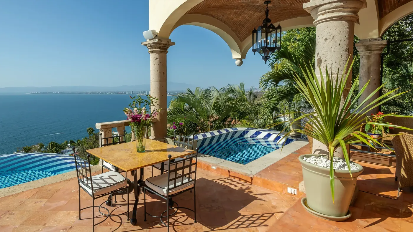 A stone-columned arched terrace with a yellow dining table beside a blue-tiled pool, looking out over the bay to Puerto Vallarta, Hacienda de los Santos