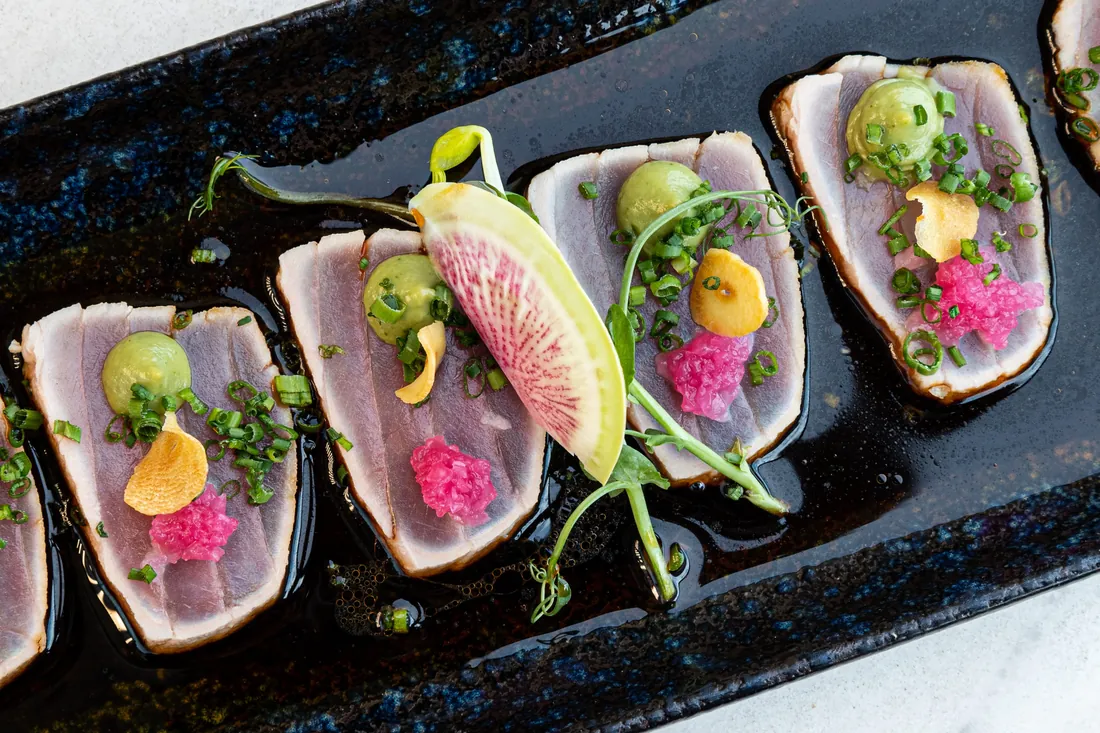 Slices of seared tuna topped with avocado cream, pink ginger and chives on a black plate