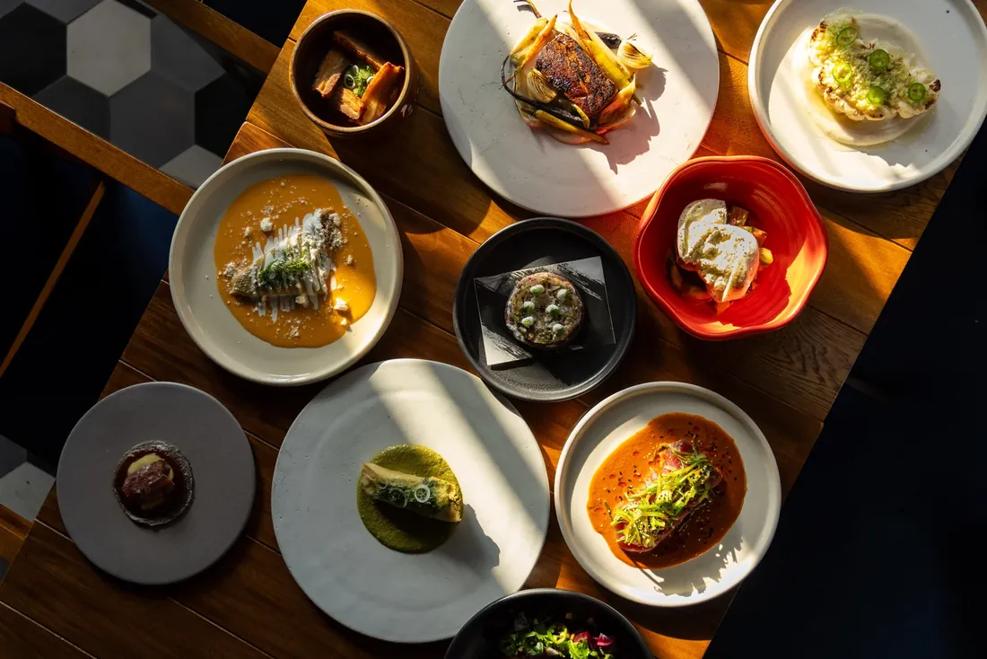 An overhead view of a wooden table covered with small plates of food in dappled sunlight