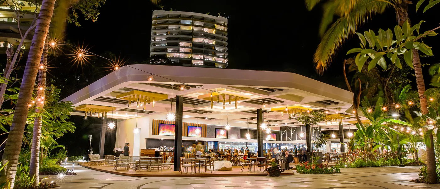 A lit open-sided restaurant pavilion among palm trees and string lights at night, with a curved hotel tower behind