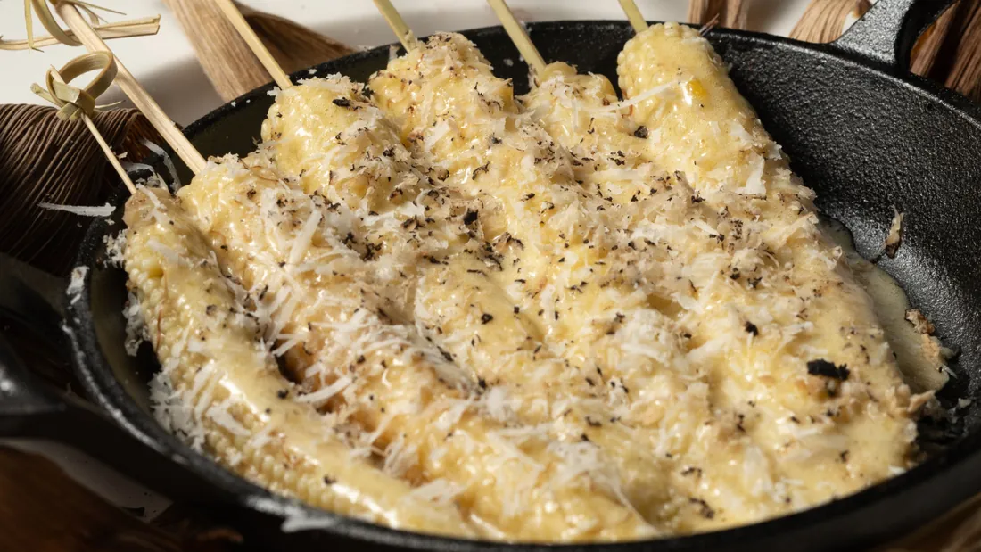 Skewers of grilled baby corn covered in grated cheese in a black cast-iron pan