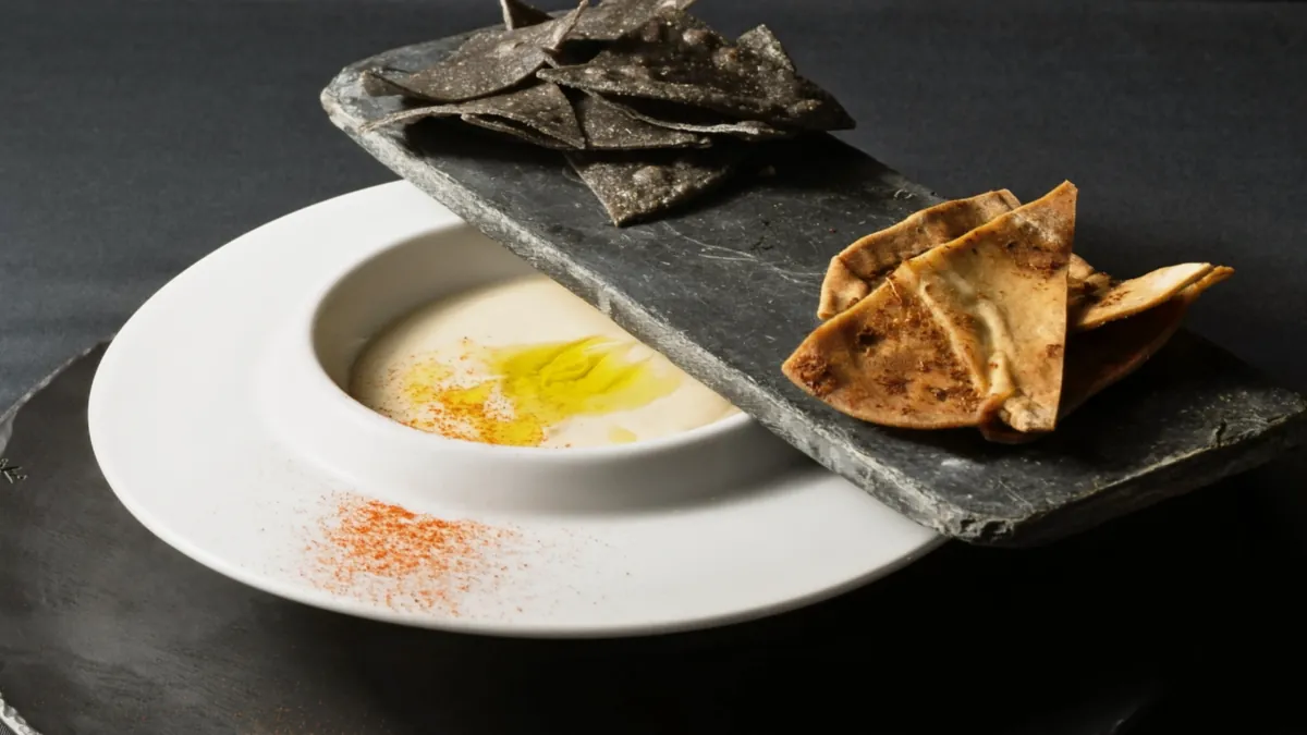 A white bowl of pale soup with olive oil and paprika beside a slate holding blue-corn chips and toasted pita
