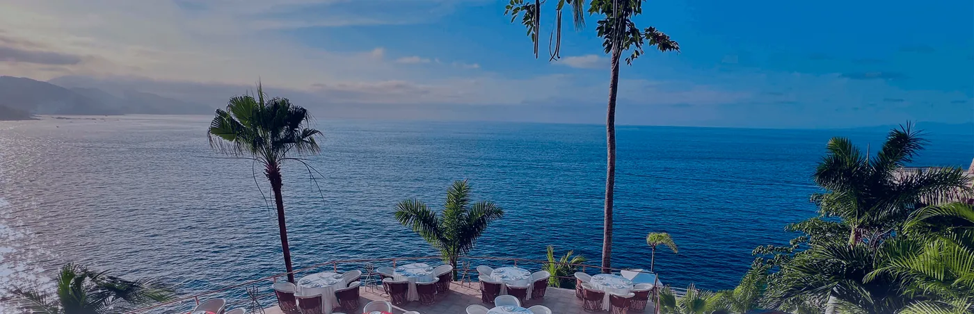 Set tables under palm trees on a stone terrace high above a blue sea, seen from above