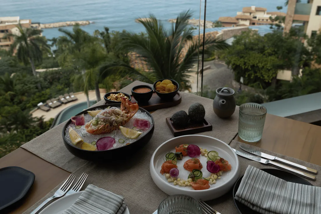 A window table set with a seafood bowl on ice, a plate of cured fish with radish and avocados, looking down over a pool, palm trees and the sea