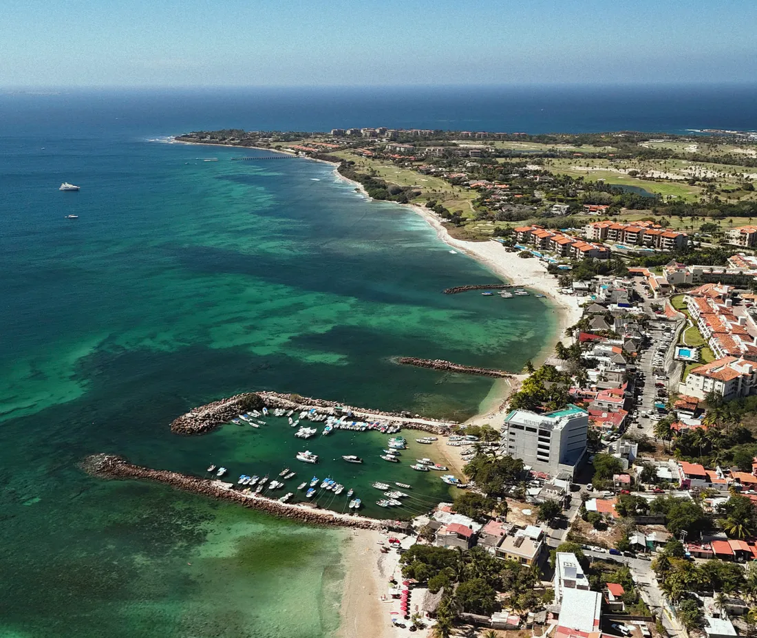 An aerial view of a small harbour and village on a green peninsula with a long beach and turquoise water