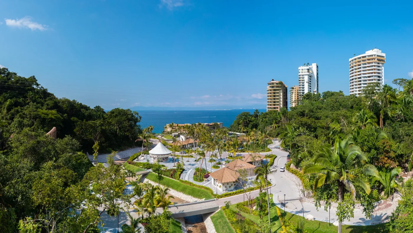 An aerial view of landscaped gardens with winding paths and a white pavilion among palm trees, with hotel towers and the sea behind