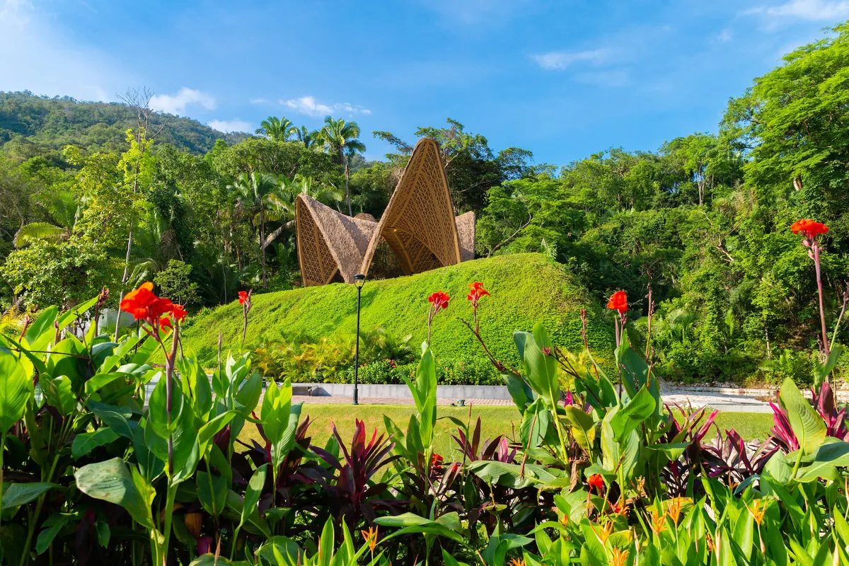 A sculptural woven-roof pavilion on a green hill behind red canna flowers and tropical plants