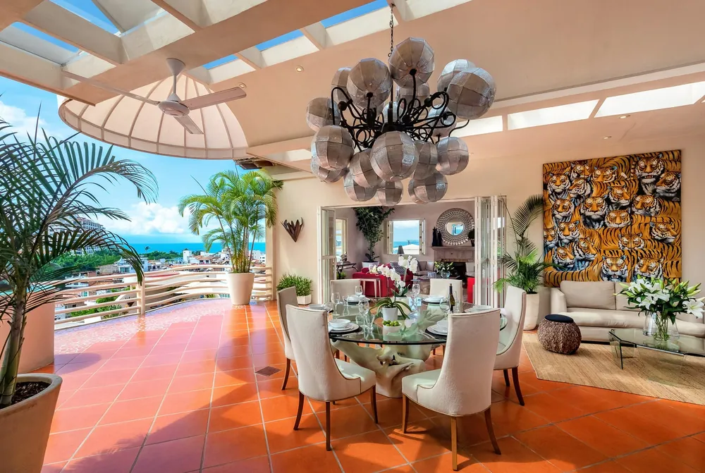 A round glass dining table with white chairs under a chandelier, open to a terrace with palms and a view over the town to the sea, Villa Tabachin, Puerto Vallarta