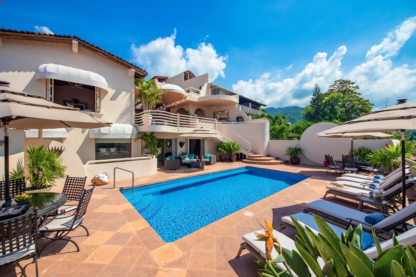 A blue pool with loungers on a tiled terrace below the white curved terraces of the villa, under a blue sky with clouds, Villa Tabachin, Puerto Vallarta