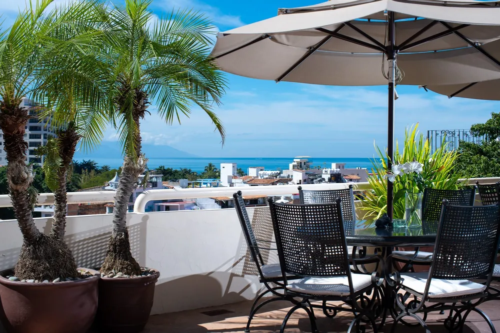 A wrought-iron table and chairs under a white umbrella beside potted palms, with white rooftops, the bay and mountains behind, Villa Tabachin, Puerto Vallarta