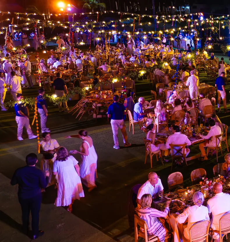 Guests dressed in white at round tables under strings of lights on a lawn at night, seen from above