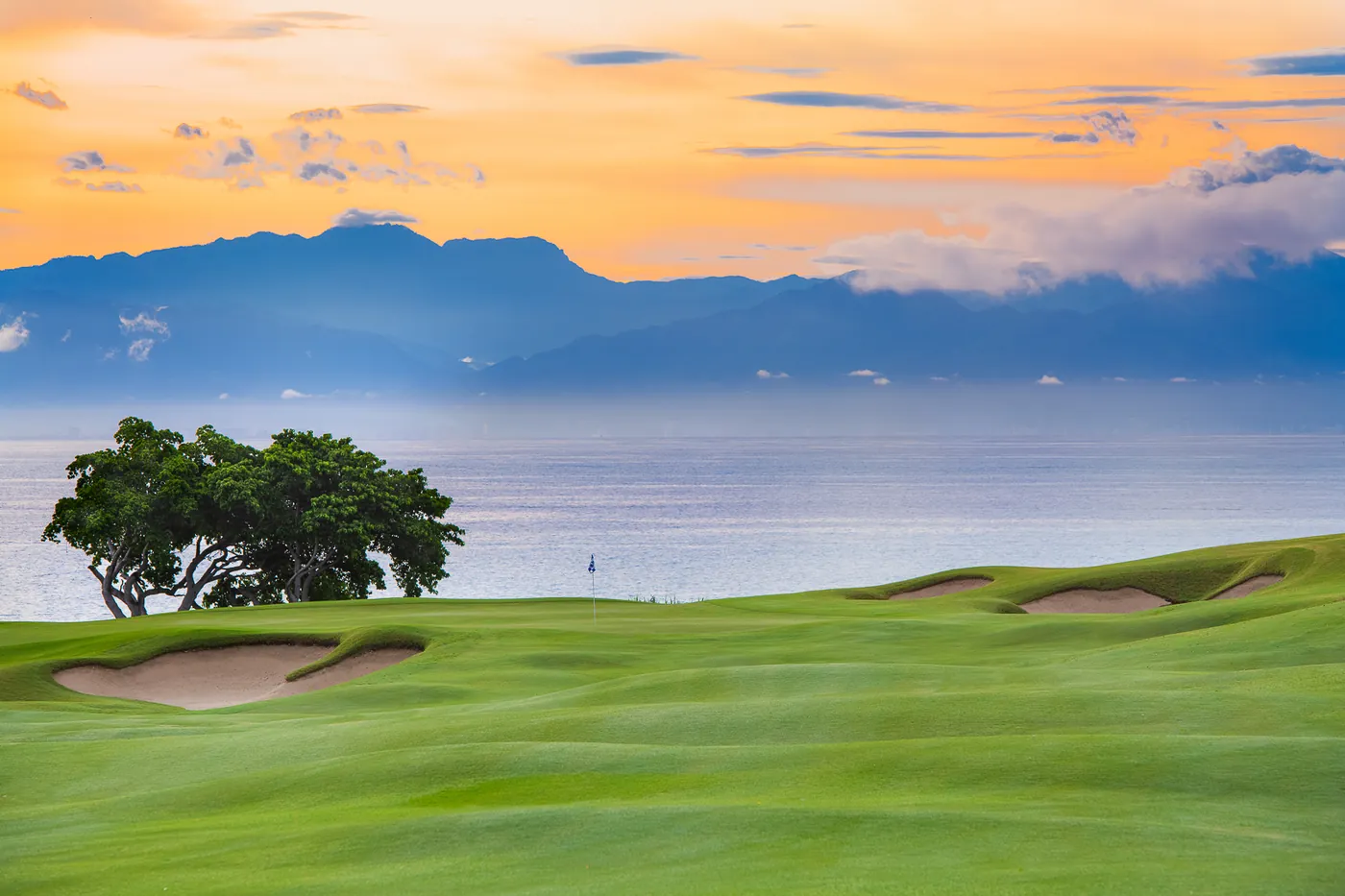 A golf green with sand bunkers and a lone tree beside the sea, with mountains and a sunset sky behind, on the Bahía course at Punta Mita