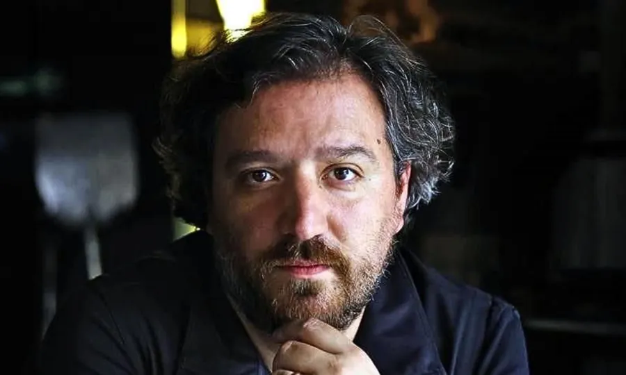 César de la Parra resting his chin on one hand, lit from the side against a dark background
