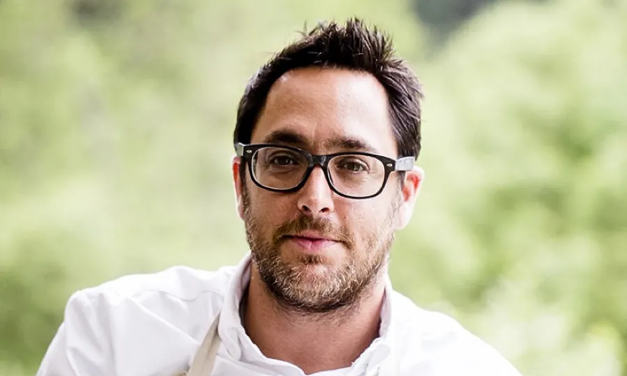Christopher Kostow in glasses and a white chef's jacket with a beige apron, trees out of focus behind him