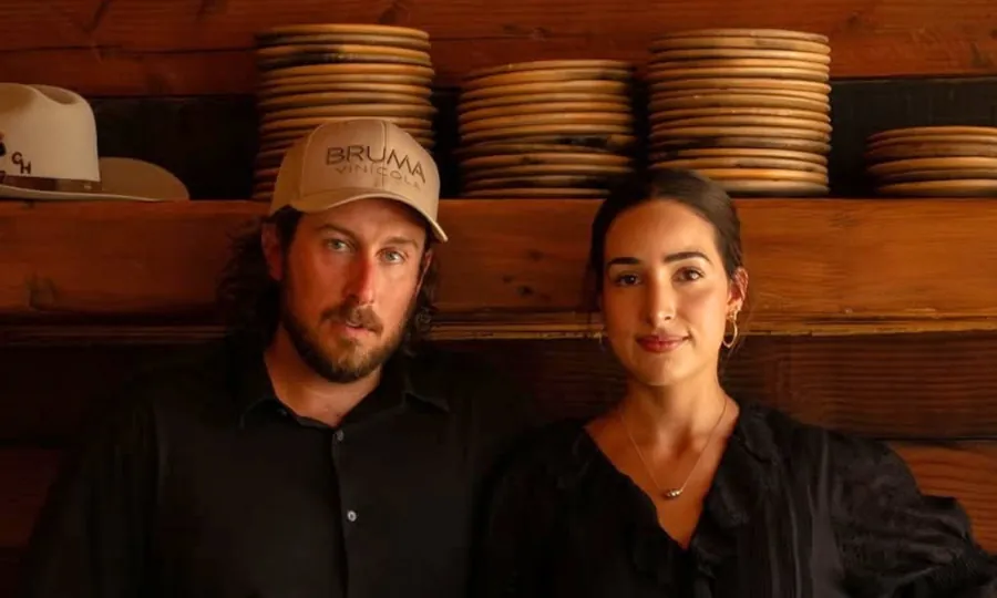 David Castro Hussong in a cap and Maribel Aldaco side by side in front of shelves stacked with clay plates